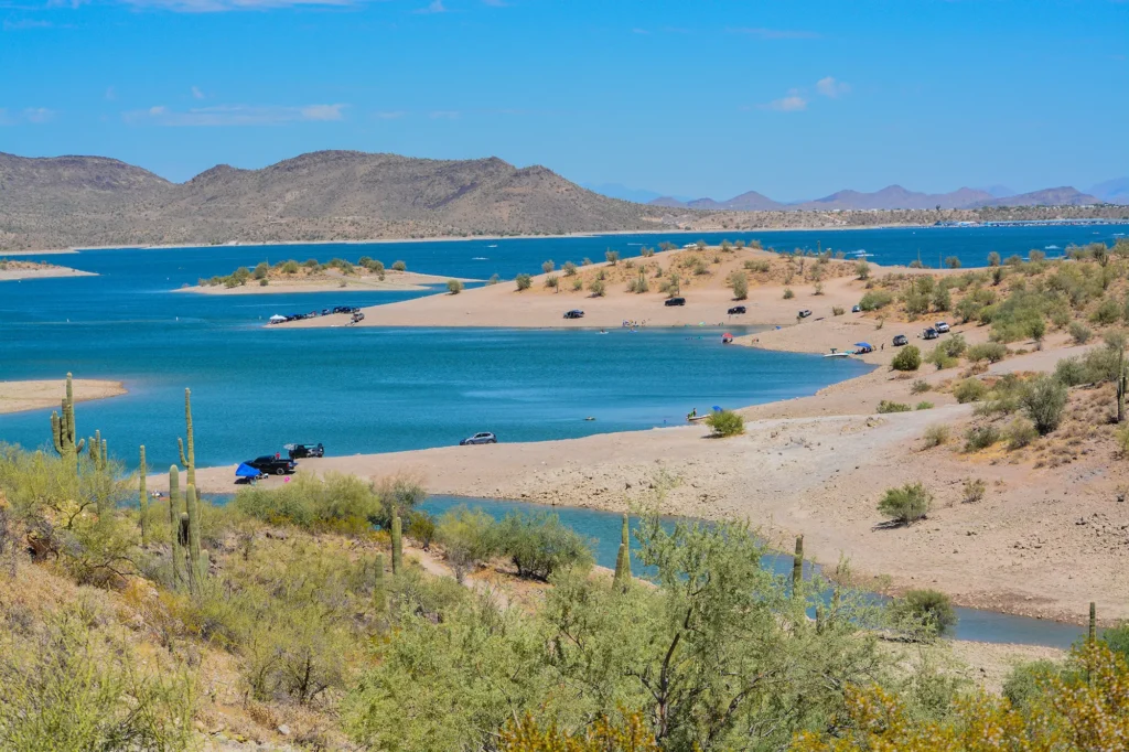 Lake Pleasant in the Sonoran Desert with sandy shores, saguaro cacti, parked vehicles, and desert mountains in the background